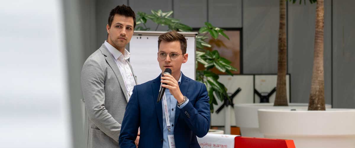 Zwei junge Männer bei einer Präsentation – einer spricht ins Mikrofon, der andere hört aufmerksam zu.